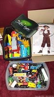 Box with assorted colorful Lego bricks inside an Alice Edge tin and plastic container, along with more toy trucks and train cars.