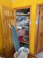 View of closet door open showing an ironing board leaning against the door and a white plastic laundry basket in front. Closet shelves hold folded linens, blankets, towels, and pillows.