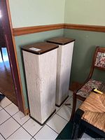 Two tall stereo speakers covered in light-colored fabric with wooden tops and bases, standing side by side in a corner.
