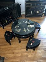 Round black lacquer tea table with glass top showing painted Asian-style bird and floral designs, displayed with four matching semi-circular stools painted with flowers, all set on hardwood floor near black lacquer furniture cabinet.