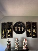 Full view of five Asian carved lacquer wall art panels mounted on a wall above three traditional Asian figurines.