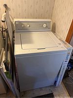 White Amana top load washing machine shown in corner of a room with box beside it.