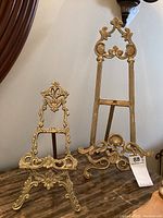 Front view of both brass easels on a marble surface showing detailed decorative patterns and relative sizes