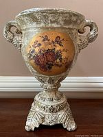 Front view of jardiniere urn showing antiqued gold floral medallion with red roses and green leaves on distressed white urn body with handles and ornate base.