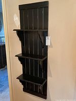 Front view of the black mounted wall shelf showing its three shelves and vertical slat back panel.