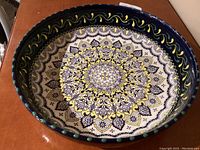 Top view showing intricate mandala floral design in blue and yellow on white background, with dark blue rim and green accents.