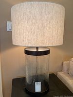 Full view of the modern glass and metal table lamp with beige fabric drum shade sitting on a black side table next to a light colored couch.