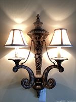 Full view of the antique style wall sconce illuminated, showing twin arms, fabric shades, and detailed floral metalwork.