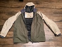 Front of jacket laid flat showing two-tone color with olive green body and beige sleeves and hood, zipped open to show inner lining.