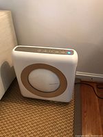 White Conway air purifier with beige ring and control panel on top with buttons and blue light on, placed on carpeted floor beside wall.
