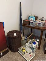 Photo shows a large dark brown wicker hamper with lid next to a grey bucket containing a mop and broom. Several plastic containers with cleaning and household items are placed beside and on a wooden table.