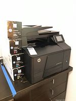 Black HP LaserJet Pro 200 Color MFP M27nw printer with four toner cartridge boxes stacked next to it.