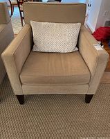 Front view of the beige upholstered armchair with white geometric accent pillow on a textured rug.