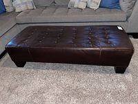 Full view of the rectangular dark brown leather bench in front of a grey sofa, showing button tufting and wooden legs.