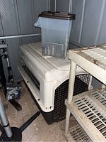 Photo showing PetMate dog kennel with a plastic food container placed on top, situated next to a metal shelving unit in a storage area, clear image of the kennel's side vents and door.