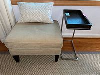 Beige hassock with matching accent pillow next to black tray side table with chrome frame on carpeted floor.