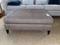 Front view of rectangular brown fabric ottoman hassock showing stained surface and tapered wooden legs