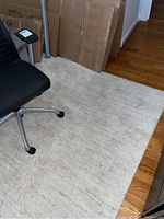 Photo showing the full rug partially under office chair and cardboard boxes, light beige with subtle texture.