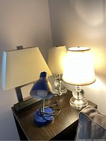 View of all four lamps grouped on a wooden table including two glass base lamps, one blue metal base lamp, and one rectangular metal base lamp with shades.