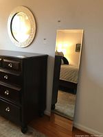 Full-length rectangular mirror leaning against wall next to dresser, showing room reflections.