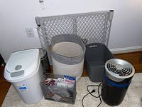 Wide shot showing all items in the lot including HoMedics air purifier, plastic baby gate, three trash cans (plastic, metal, woven), metal mesh file holder with office supplies, and wall hangers.