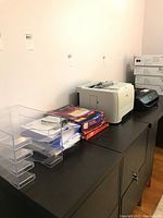 Wide view of clear plastic file organizer, file folders, copy paper stacks, HP LaserJet printer, and HP Envy printer on a black table against a wall.