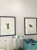 Two framed dog silhouette prints hanging side by side on a white wall above a surface with a plant and a decorative blue and green basket.