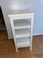 Front view of white wooden side table showing the square top and three shelves, scratches visible on the surface.