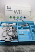 Original Nintendo Wii console packaging with Wii Sports game, two white Wii remotes, sensor bar, and cables inside the box and packaging inserts.