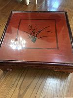 Square wooden floor table with reddish lacquer finish and hand-painted floral motif on top surface, showing one damaged leg.