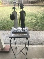 Full view of the brass metal sculpture man and woman on a decorative metal and tile side table, showing the figures from front and side angle with outdoor yard background.