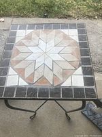 Square outdoor foldable table with patterned tile top and black wrought iron legs on a concrete surface with grass background.
