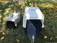 Photo of two pet crates on grass. Larger crate is blue and gray with open metal door, smaller crate is black and gray with plastic front, handle on the large crate missing.