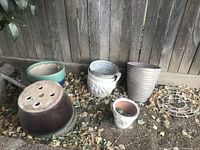 Various plant pots of different sizes and colors arranged on the ground near a wooden fence with fallen leaves.