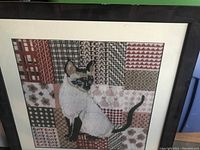 Framed needlepoint tapestry of Siamese cat with patchwork background showing embroidery detail.