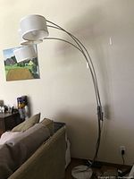 Full view of the Uttermost Bradenton floor lamp by the wall beside a couch, showing three arched metal arms and drum shades, with marble base on floor.