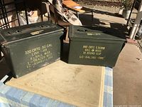 Two vintage military green ammo boxes side by side on a table, showing their yellow stenciled label text distinctly.