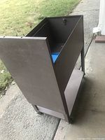 Side and partial back view of vintage brown metal file cabinet on wheels, showing tall rectangular shape with lower shelf and top open compartment with lock.