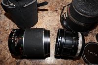 Top view of two vintage camera lenses showing textured grip, focus rings, distance scales, and lens caps. Two leather carrying cases visible.