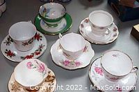 Six tea cups with matching saucers arranged on a table showing different floral patterns and a green cup with visible chip.