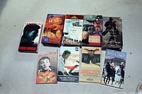 Top-down view of nine VHS movie tapes arranged in two rows on a gray concrete floor. Titles and cover art clearly visible. VHS boxes shown face up.