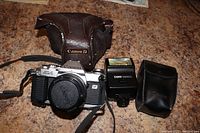 Canon AL-1 camera with 50mm lens attached, skylight filter, lens cap, Canon 166A Speedlite flash, soft flash case, and worn brown leather Canon carrying case arranged on wood surface.