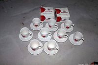 Nine white ceramic coffee cups with red Grand Marnier logo and seal on the front, arranged with matching saucers.
