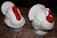 Front and angled view of two ceramic turkey serving dishes with white bases and detailed turkey lids with red wattles and snoods