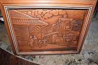 Full view of the wooden bas relief carving showing a train station scene with a locomotive and people on the platform, mounted in a wooden frame.