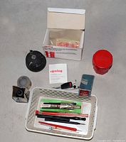 Top-down view of various vintage pen and drafting supplies arranged on a floor including Rotring pen cleaner, ink bottles, pencil leads, scribber, and drafting tools