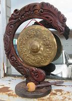 Front view of the carved wooden dragon frame holding the brass dinner gong with red hanging cord, showing the intricate dragon and floral embossing on the gong surface alongside the wooden striker with padded head on the base.