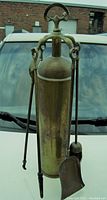 Front view of the antique trench art shell form fireplace implement stand with attached tools, showing patina and overall shape.