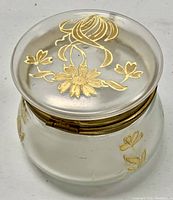 White frosted glass round dresser box with lid showing gold enamel floral design and metal rim with hinge on white background.