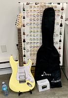 Full view of yellow Donner electric guitar on a stand with black carrying case, mini amp box, and power cord in front.